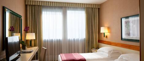 Habitación de hotel con dos camas, un escritorio con televisor, una silla, una lámpara y un cuadro en la pared.