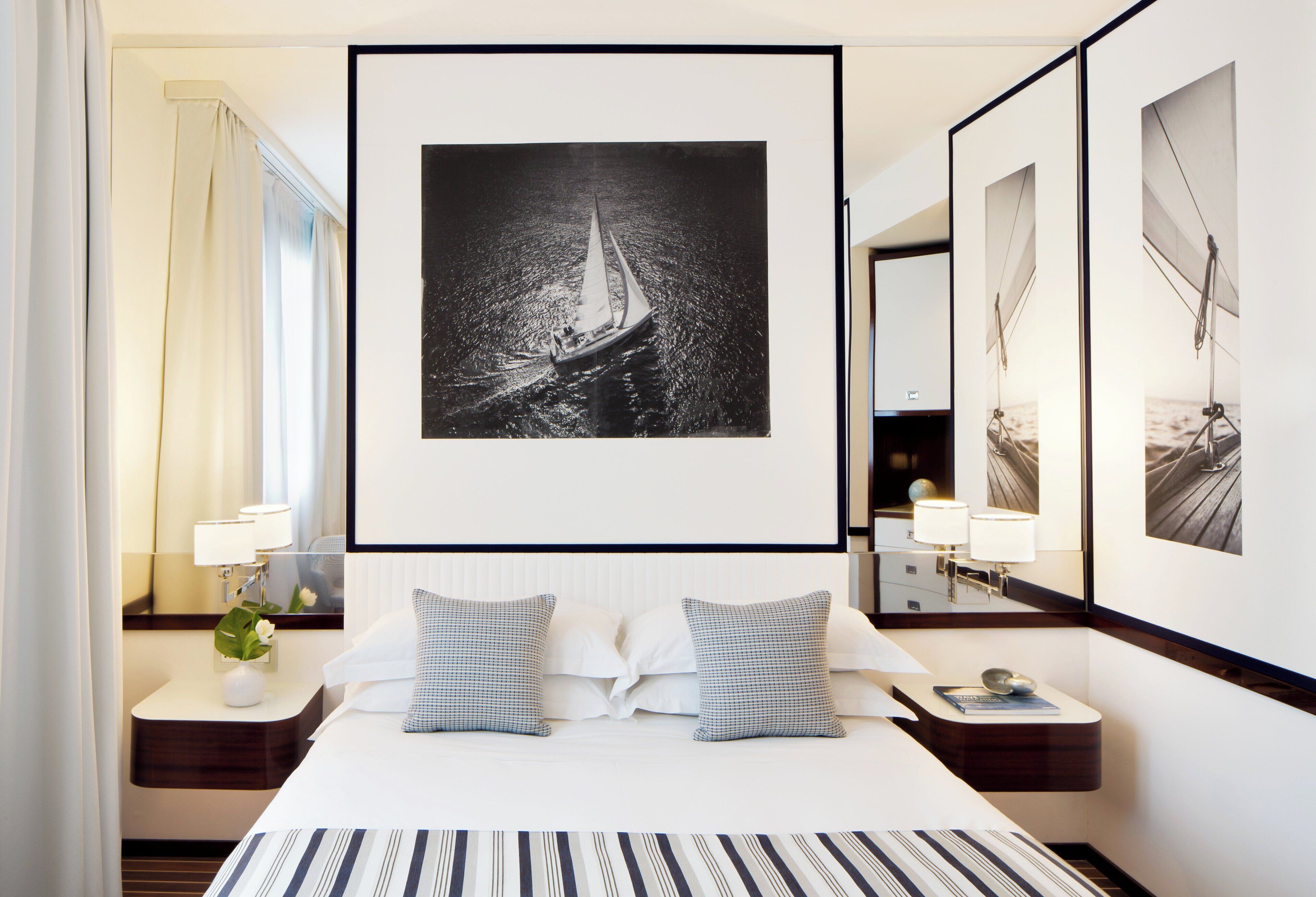 A hotel room with a large bed, two pillows, a nightstand with a lamp, a mirror, and a framed black and white photograph of a sailboat on the wall.
