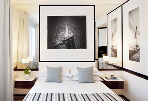 Habitación de hotel con una cama grande, dos almohadas, una mesita de noche con lámpara, un espejo y una fotografía en blanco y negro de un velero en la pared.