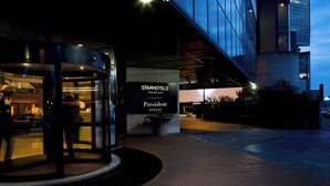Vue extérieure de l’hôtel StarHotels Premium President Genova au crépuscule, avec sa façade en verre, son entrée dotée d’une porte tournante et son hall d’entrée bien éclairé.