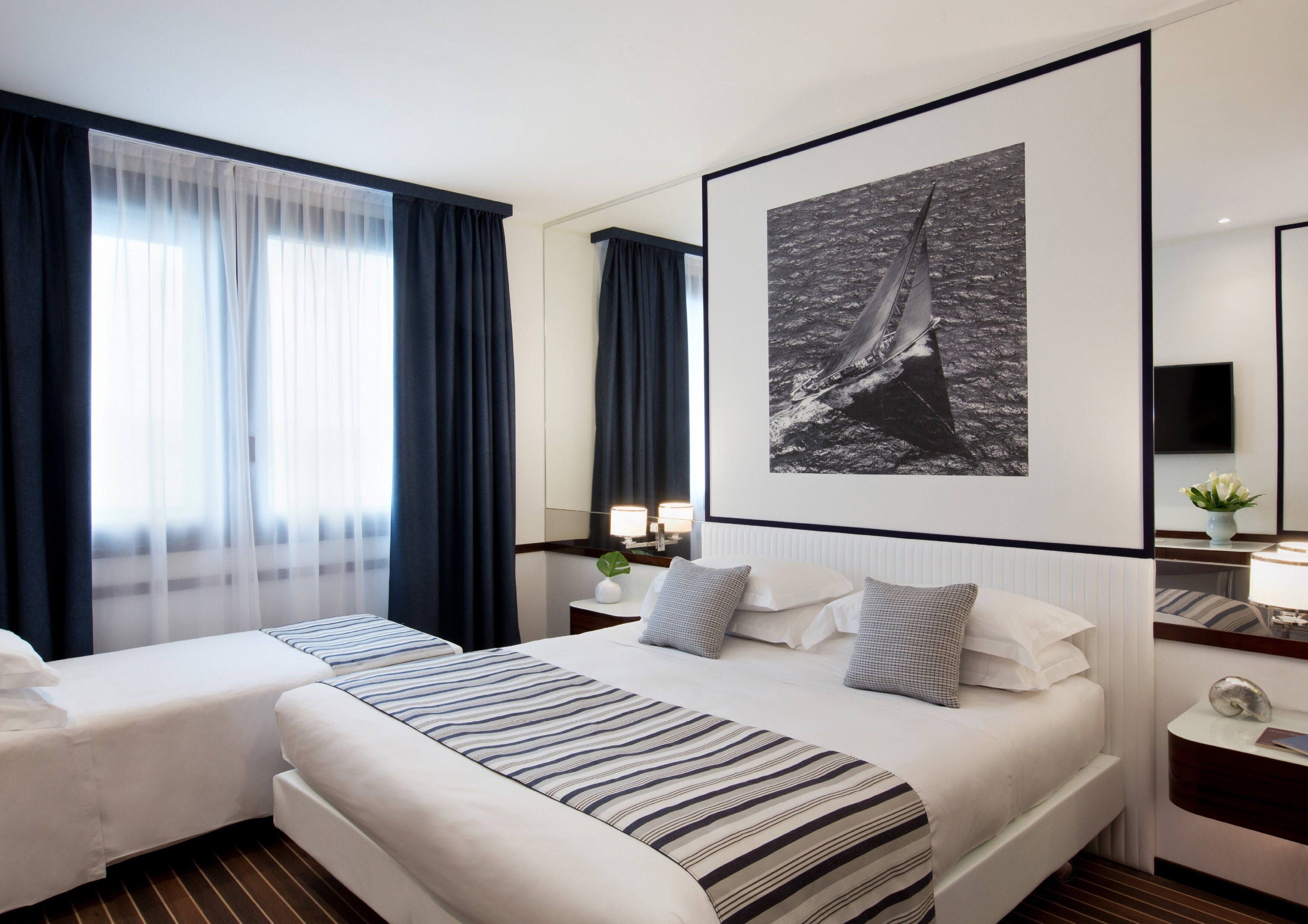 A hotel room with two beds, a large framed picture of a sailboat, a bedside table with a lamp, and a mirror on the wall.