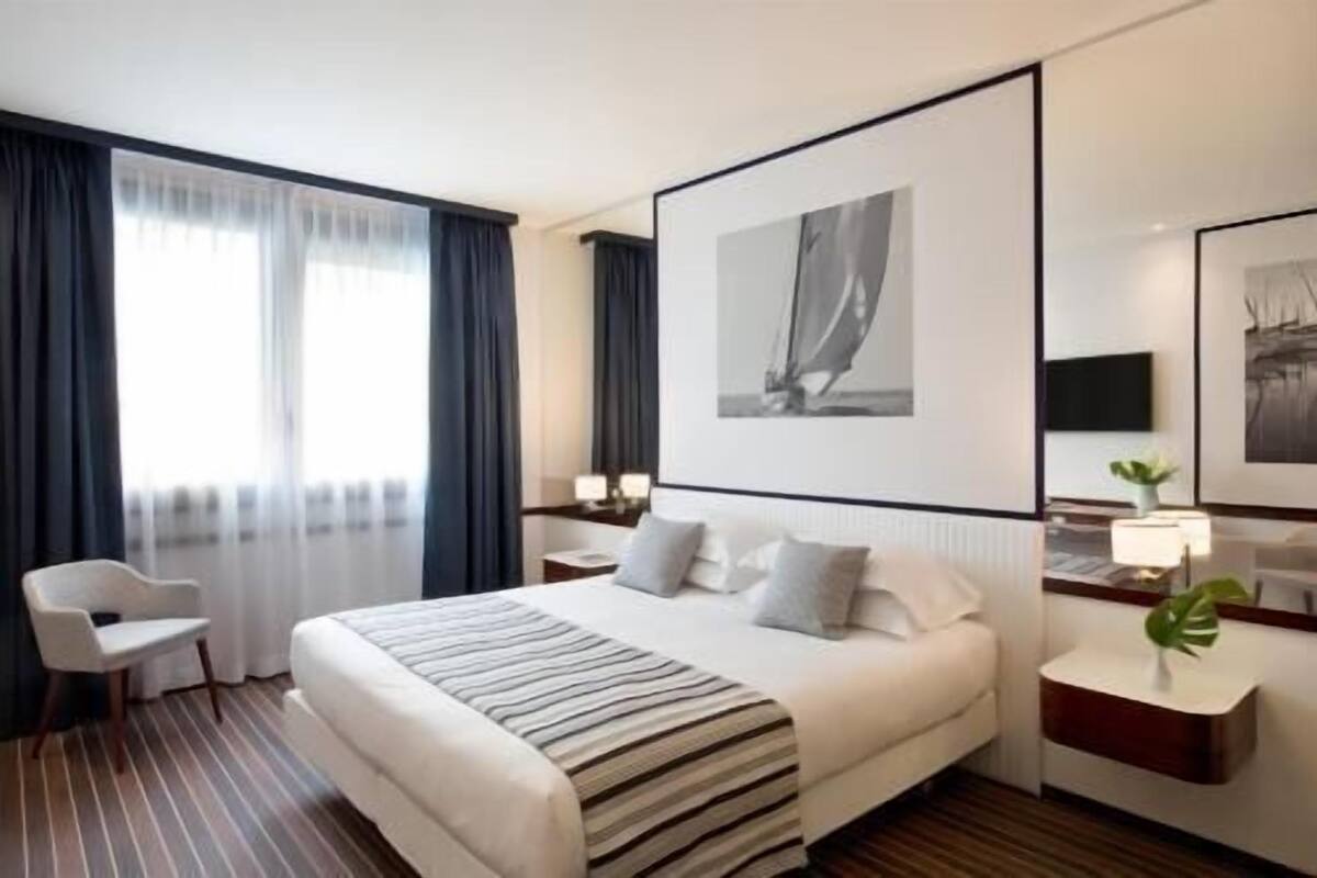 Une chambre d’hôtel avec un lit, une chaise, un bureau, une lampe et des œuvres d’art accrochées aux murs.