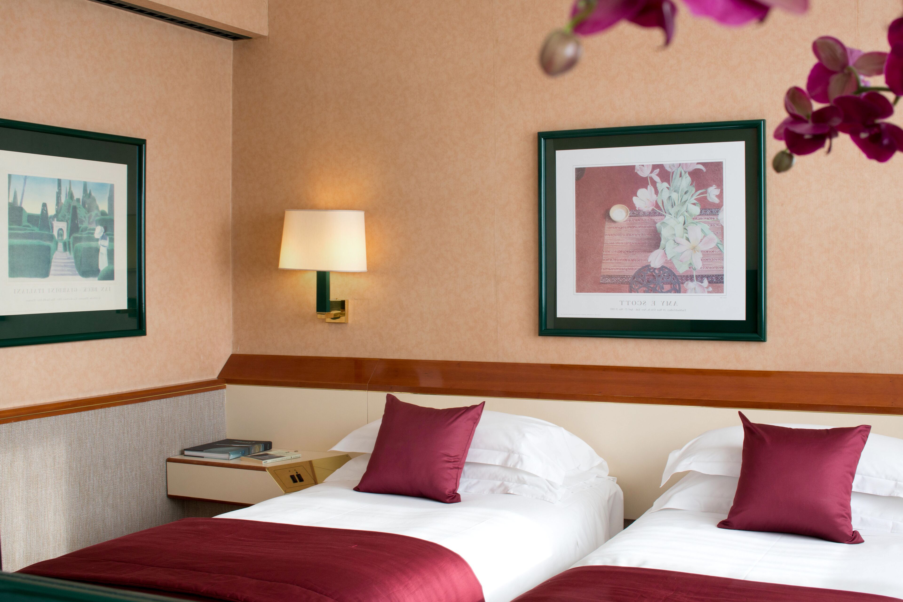 A hotel room with two beds, a nightstand, a lamp, and framed pictures on the walls.