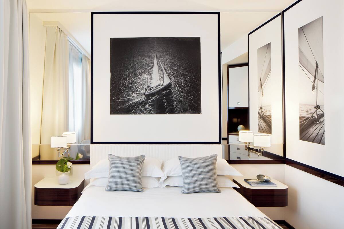 Une chambre d’hôtel avec un grand lit, deux oreillers, une table de chevet avec une lampe, un miroir et une photographie en noir et blanc encadrée représentant un voilier, accrochée au mur.
