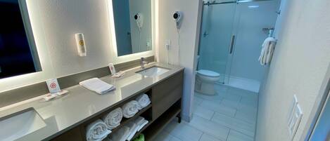 Un baño moderno con un espejo grande, lavamanos dobles, cabina de ducha de vidrio y toallas ordenadas en estantes.