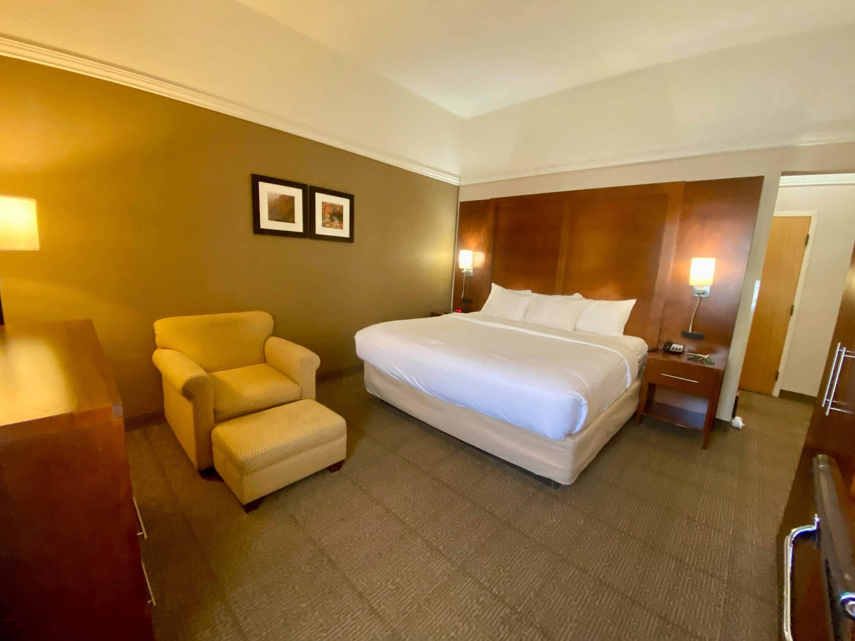 A hotel room with a large bed, a chair, a desk, and two framed pictures on the wall.