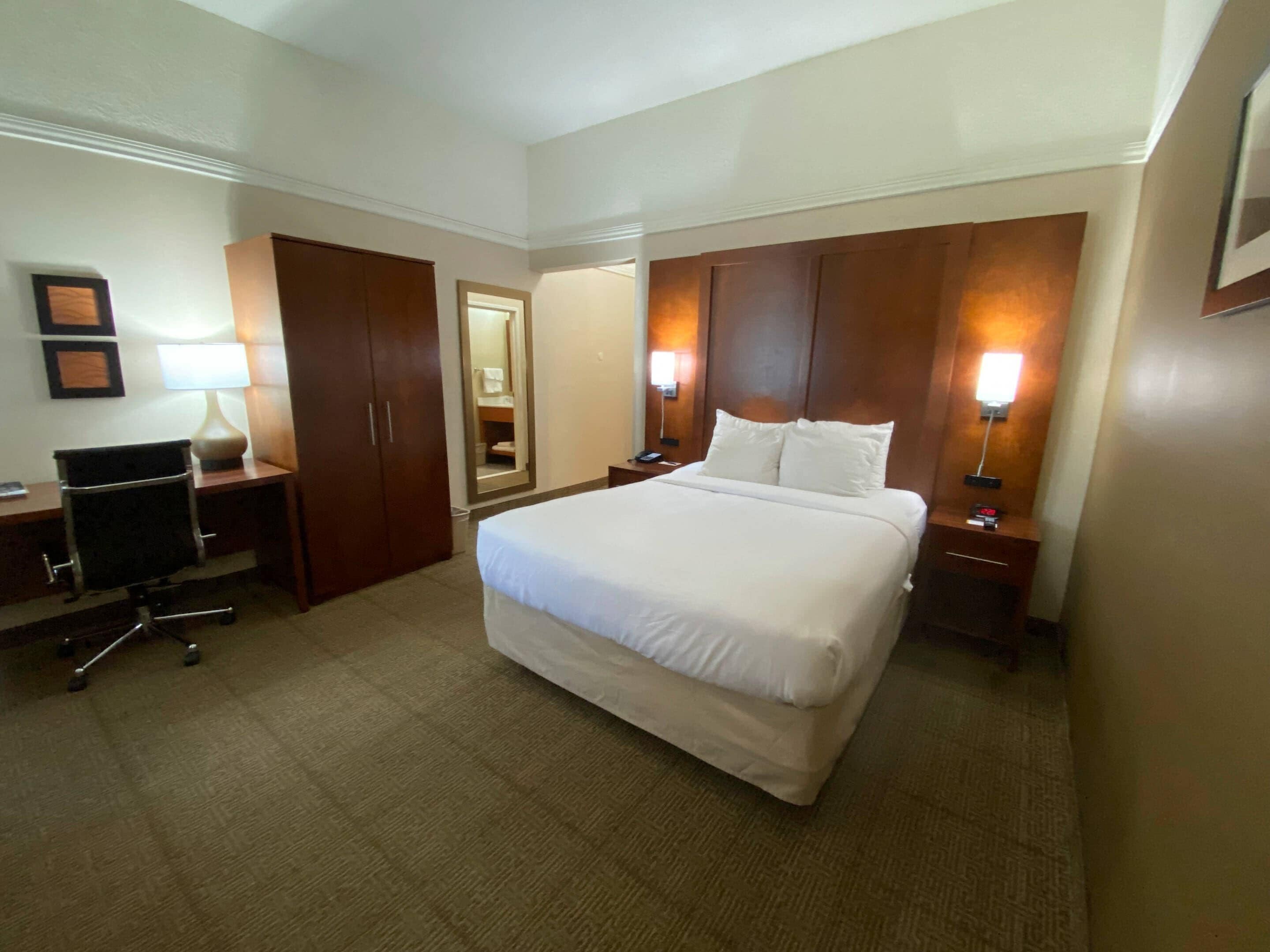 A hotel room with a large bed, a desk with a chair, a wardrobe, and two bedside lamps.