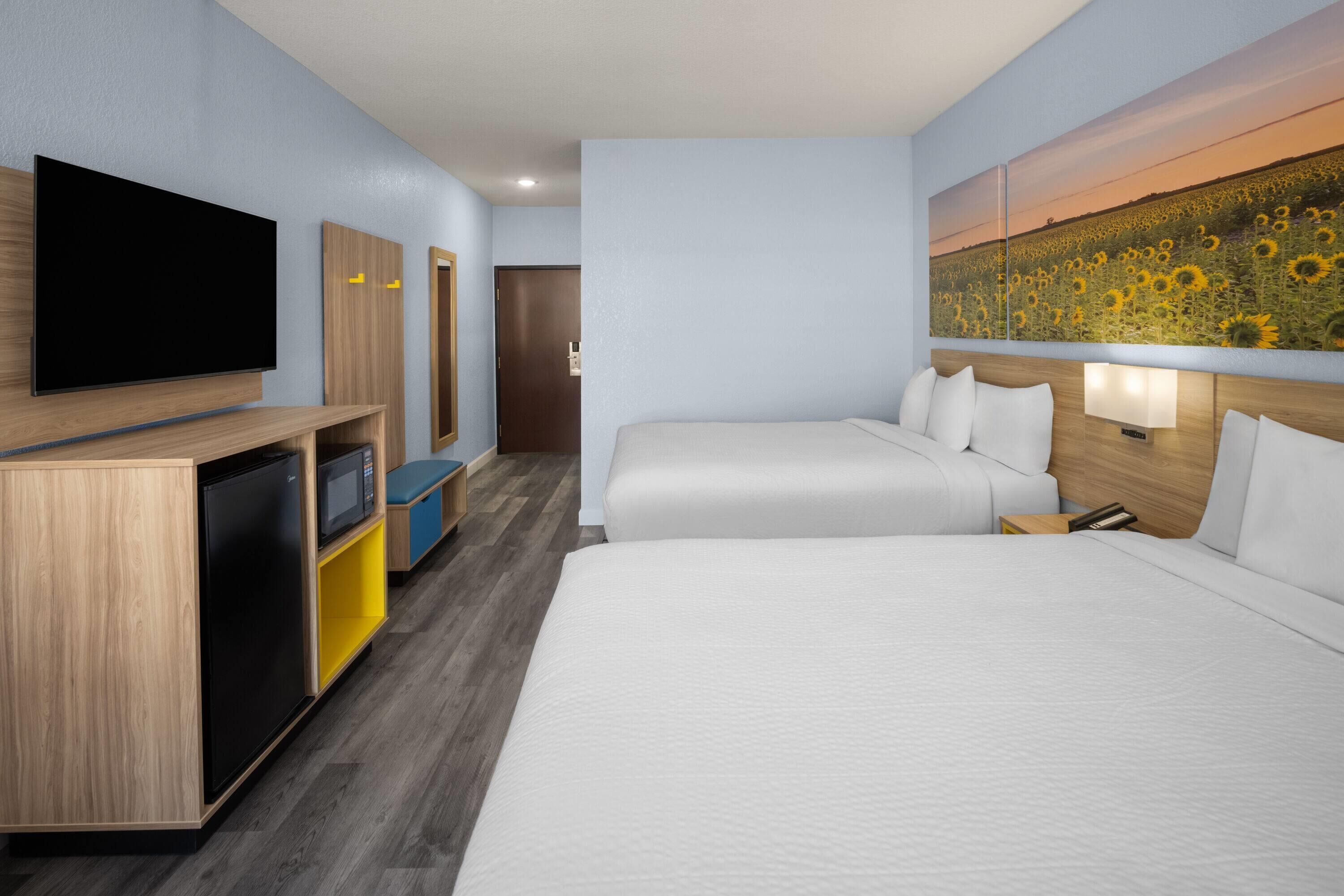 A hotel room with a TV, a desk with a microwave, two beds, and a painting of sunflowers.