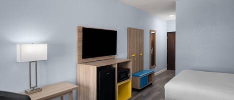 A hotel room with a bed, a desk with a lamp, a TV, a mini-fridge, and a wardrobe.