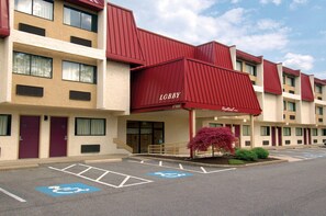 Un hôtel à plusieurs étages, doté d’une marquise rouge sur laquelle on peut lire les mots « Réception » et « Red Roof Inn ». Des places de stationnement sont prévues, et elles sont signalées par des marquages bleus indiquant qu’elles sont accessibles aux personnes handicapées.
