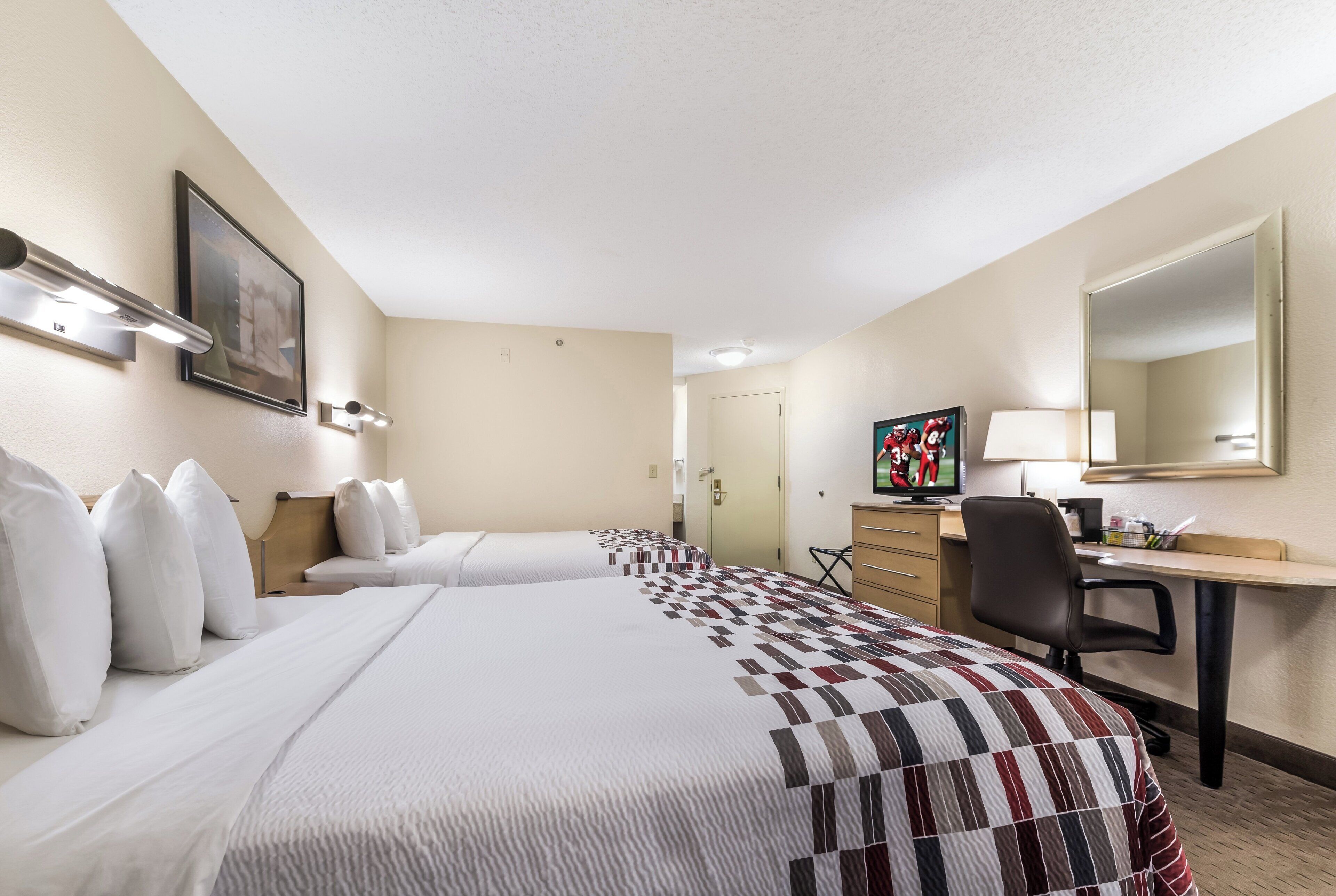 Ein Hotelzimmer mit zwei Betten, einem Schreibtisch, einem Stuhl, einem Fernseher und einem Bild an der Wand.