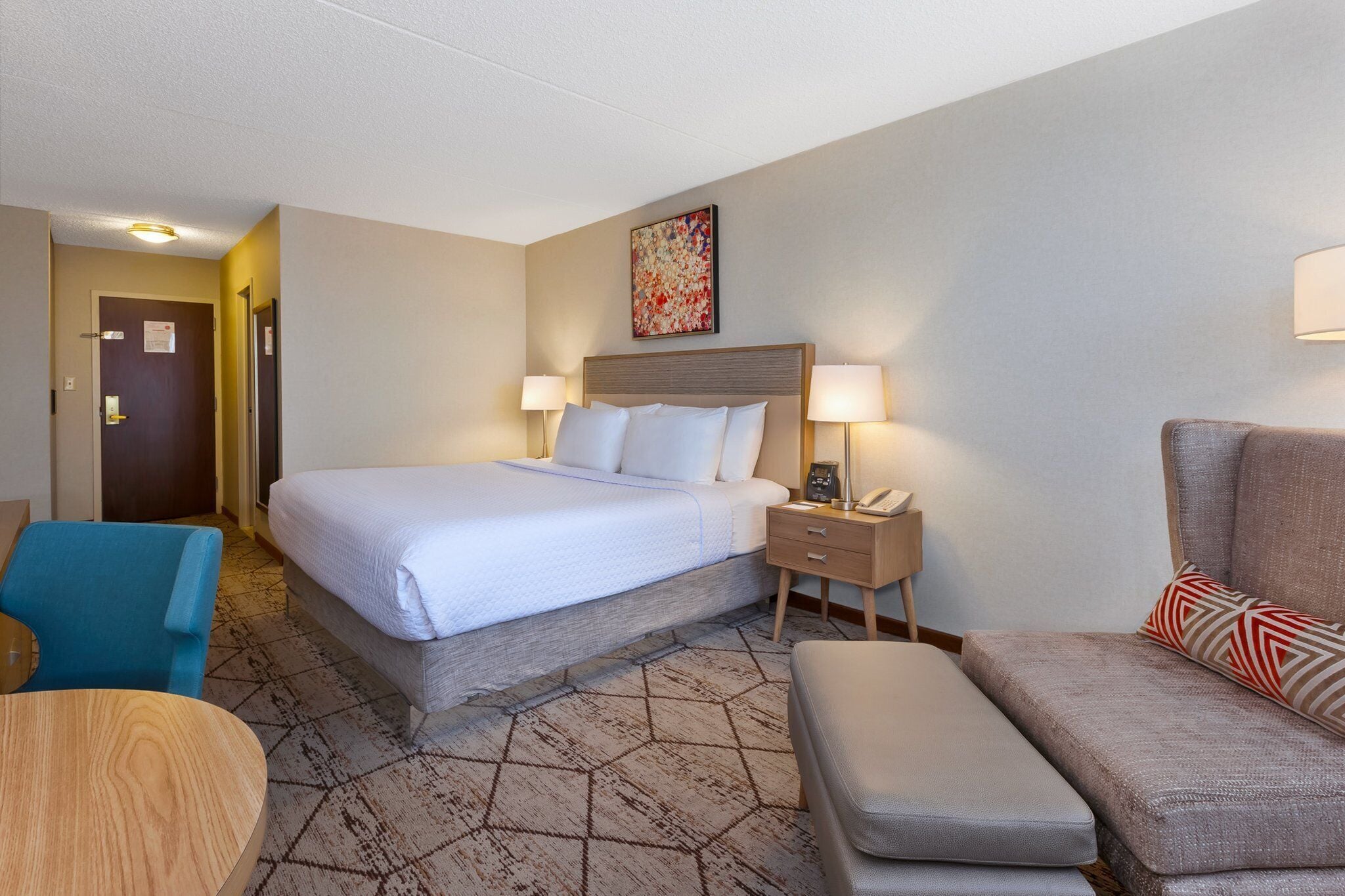A hotel room with a bed, bedside lamps, a desk, a chair, and a sofa.