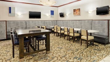 A conference room with a long table and multiple chairs, equipped with a flat-screen TV and wall-mounted speakers.
