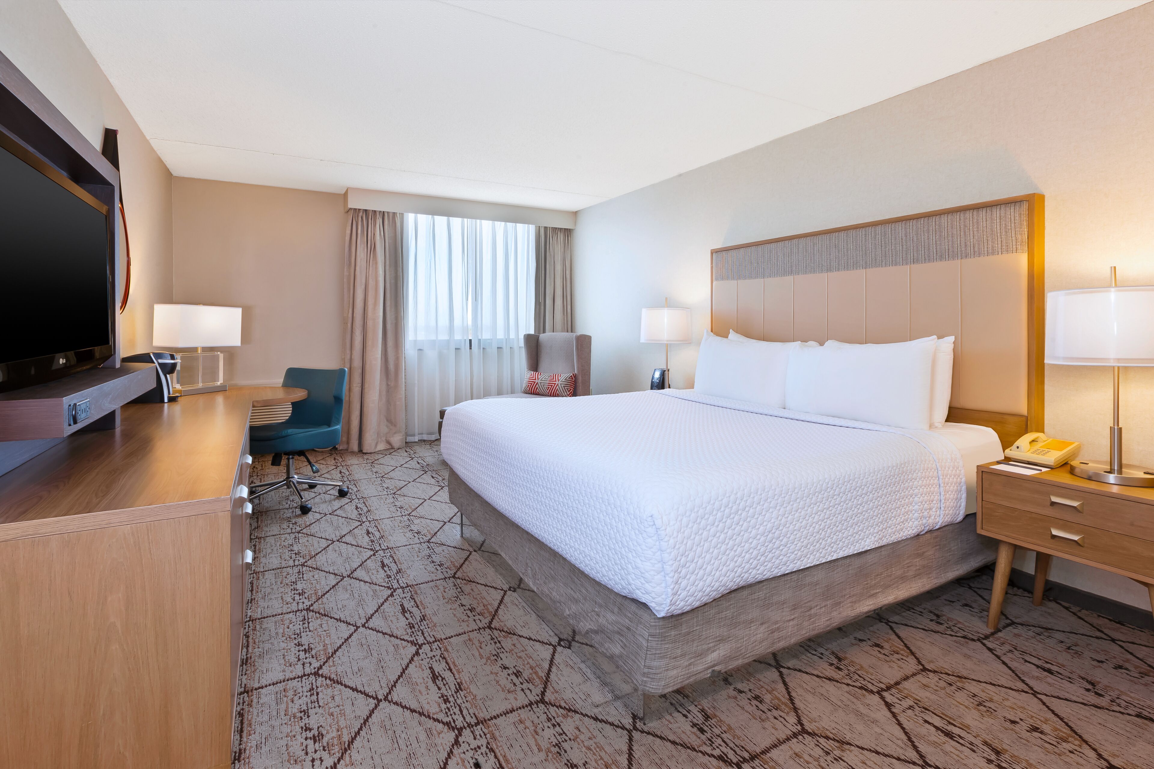 A hotel room with a large bed, a desk with a flat-screen TV, a chair, and a lamp.