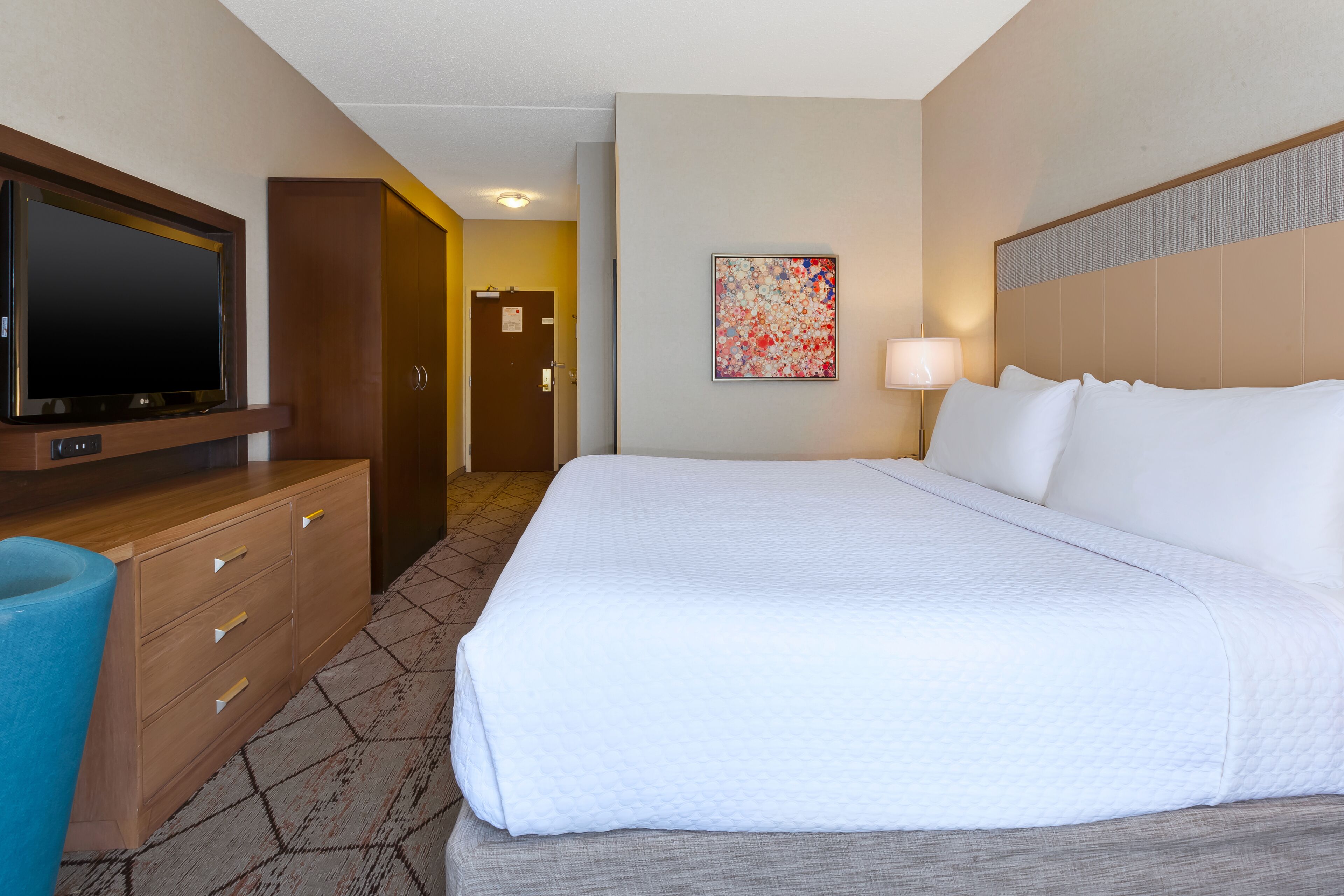 Une chambre d’hôtel avec un grand lit, un téléviseur à écran plat fixé sur une commode en bois, un tableau au mur et une porte donnant sur une autre pièce.