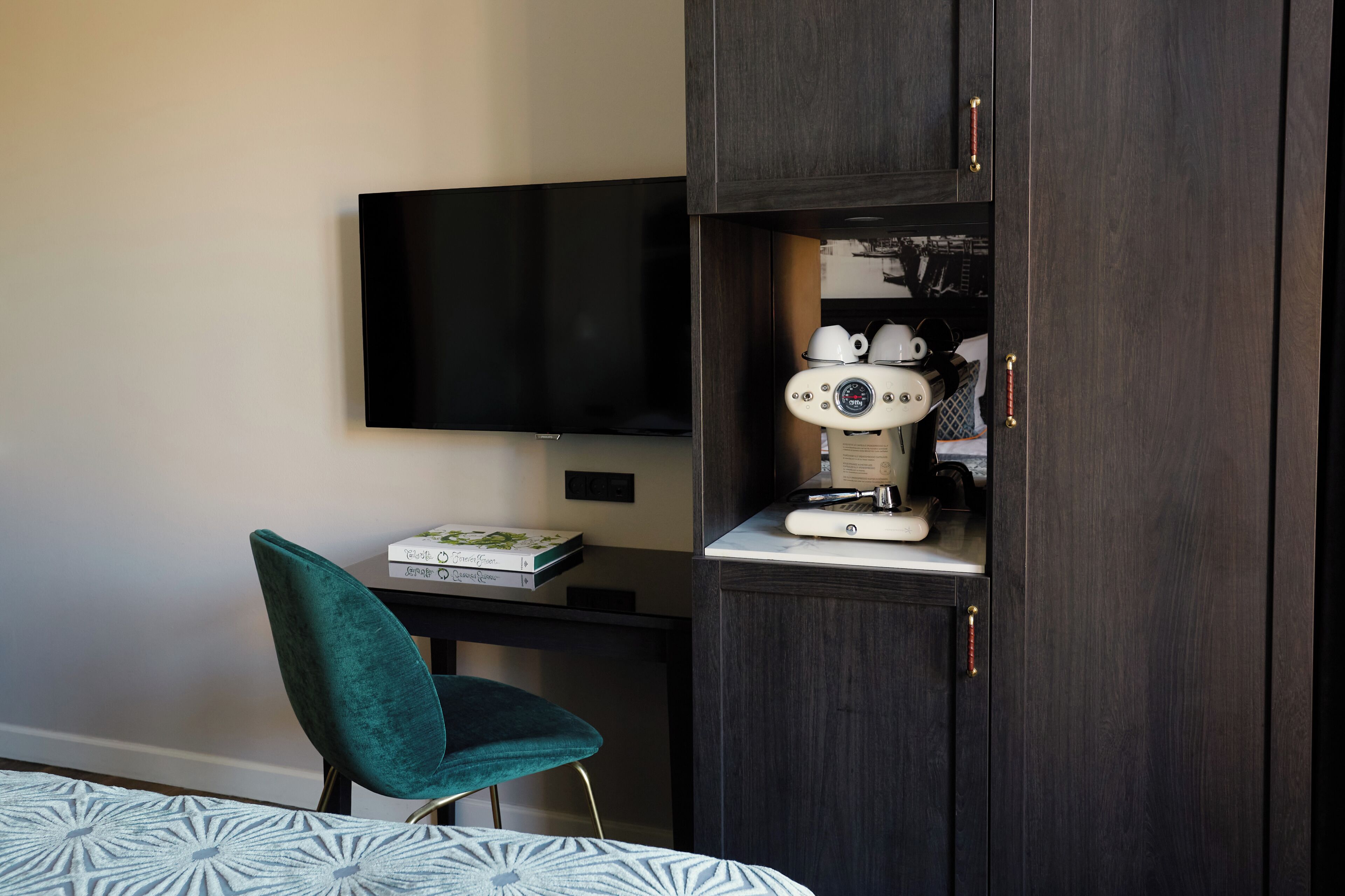 Una habitación con un armario de madera oscura, un televisor de pantalla plana en la pared, una silla color turquesa y una cama con colcha estampada.