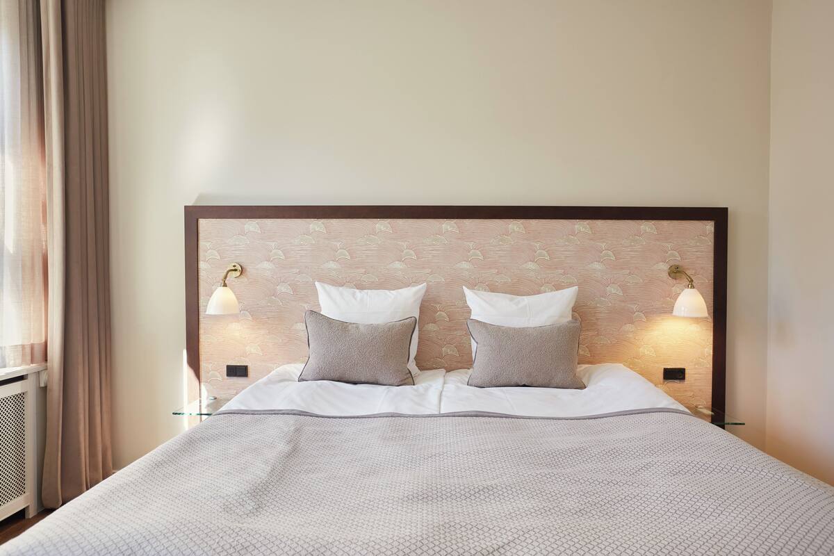 Una cama bien hecha con un cabecero estampado, dos lámparas de noche y una ventana con cortinas.