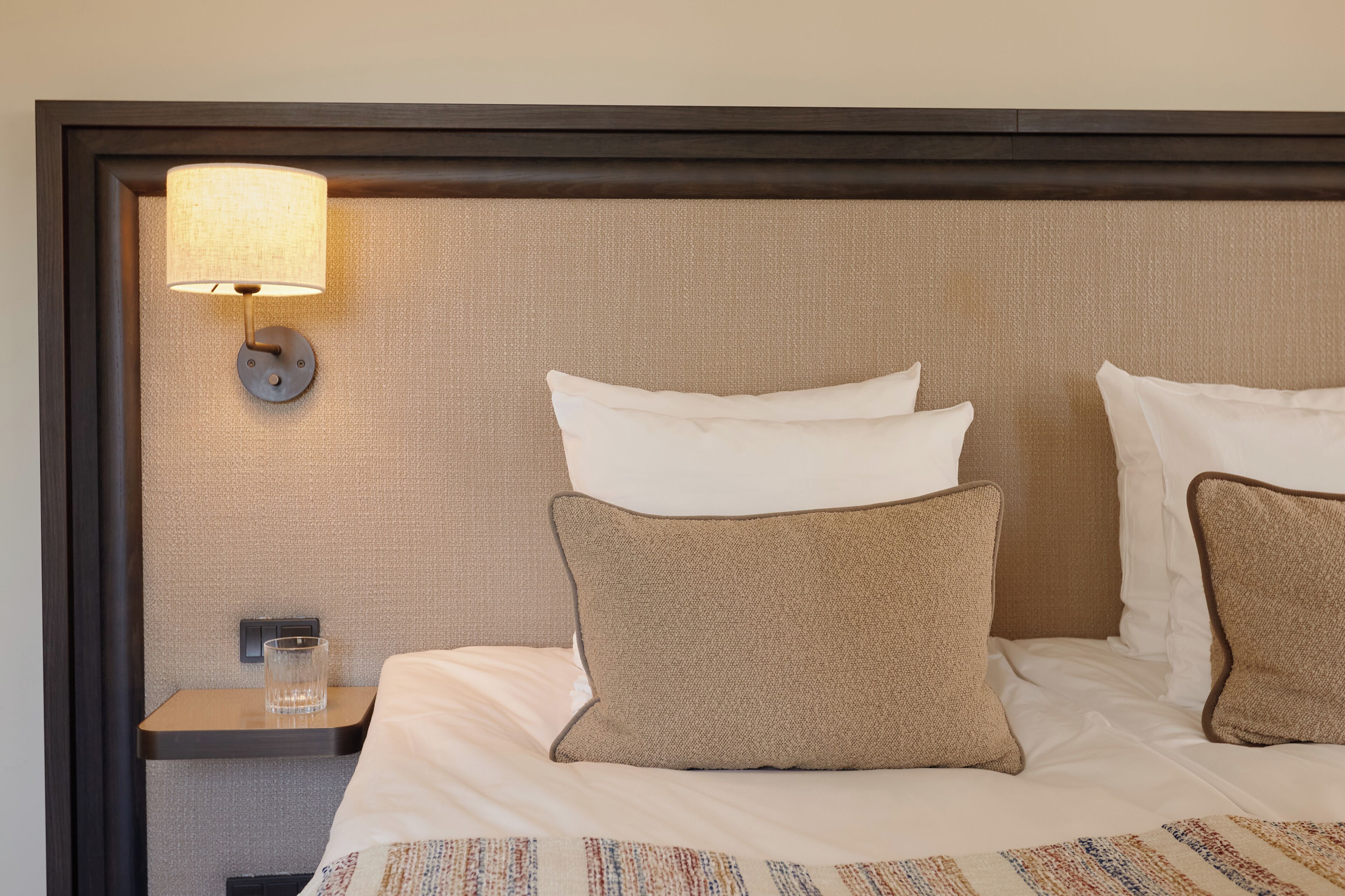 Una cama bien hecha con almohadas blancas y marrones, una mesita de noche con un vaso y una lámpara fijada a la pared.