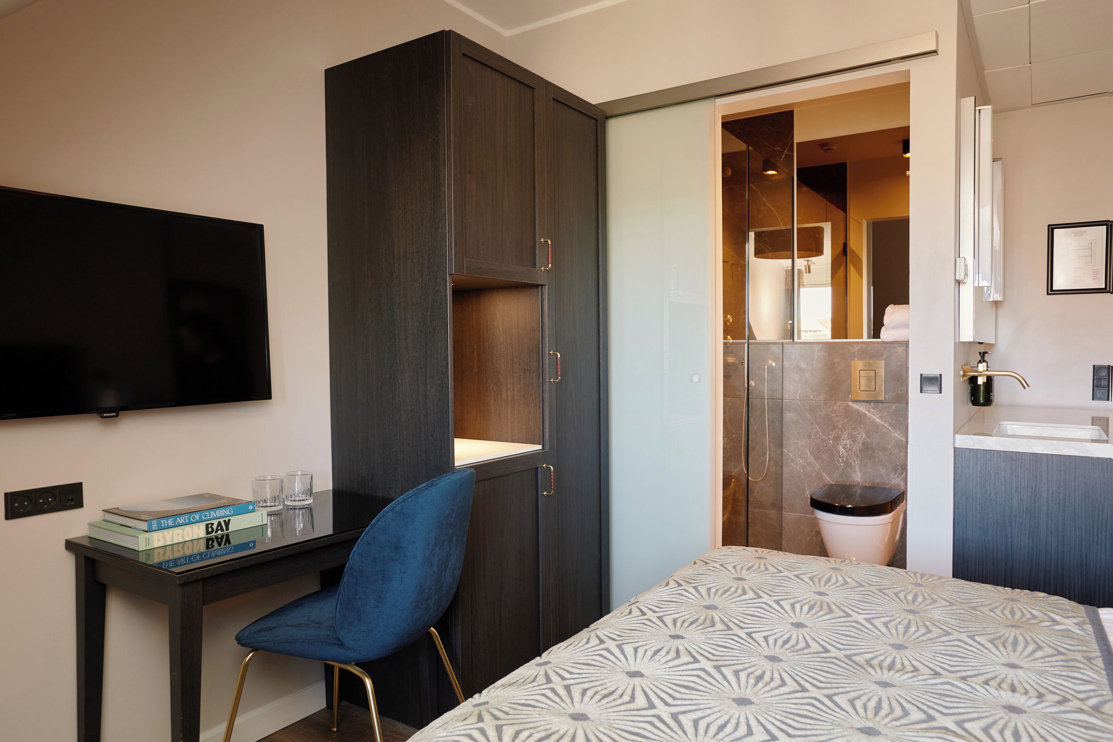 A hotel room with a bed, a desk with a chair, a TV, a wardrobe, and a bathroom.
