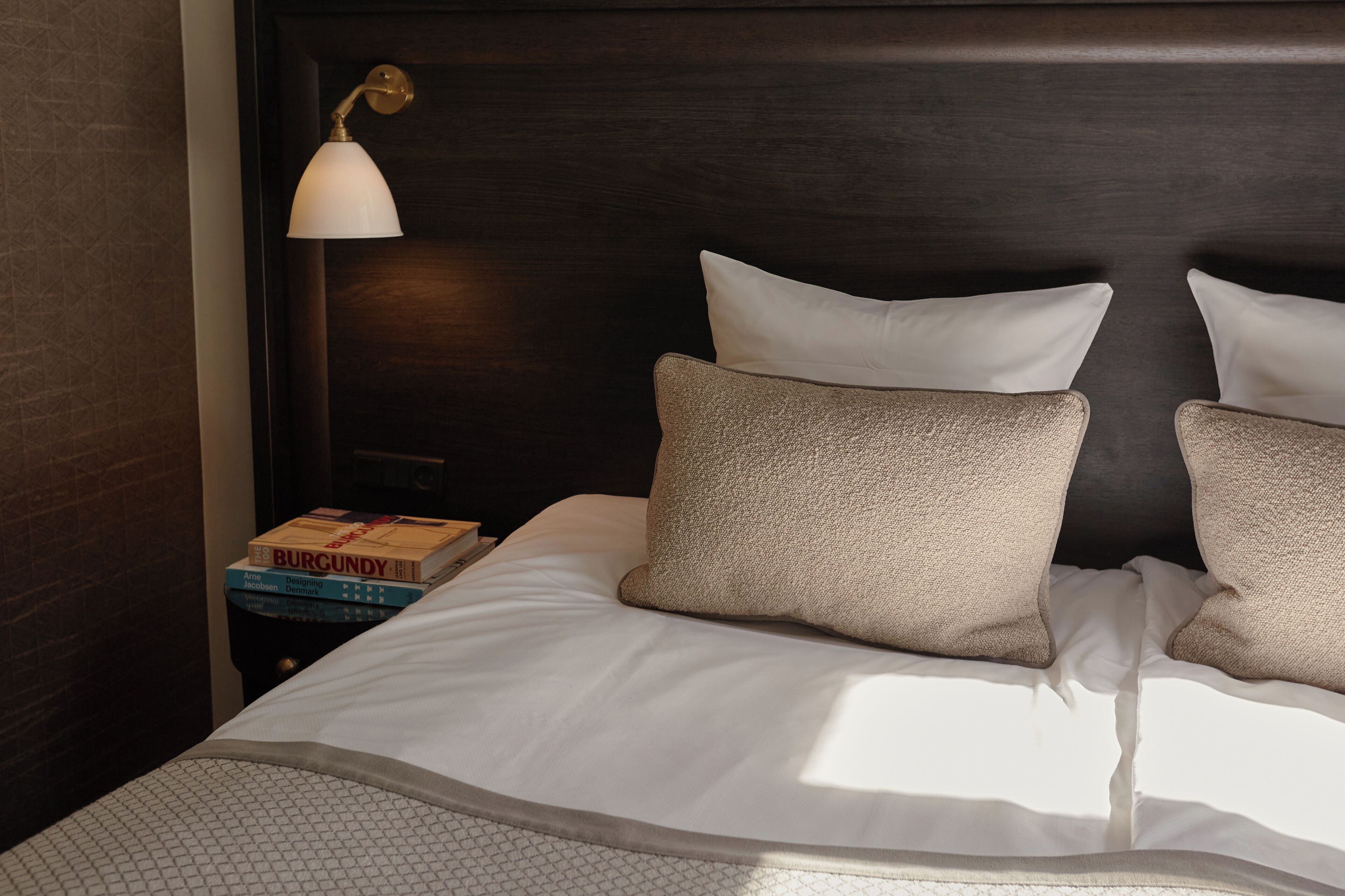 A neatly made bed with white linens and two beige pillows, a small bedside table with a lamp, and a stack of books.