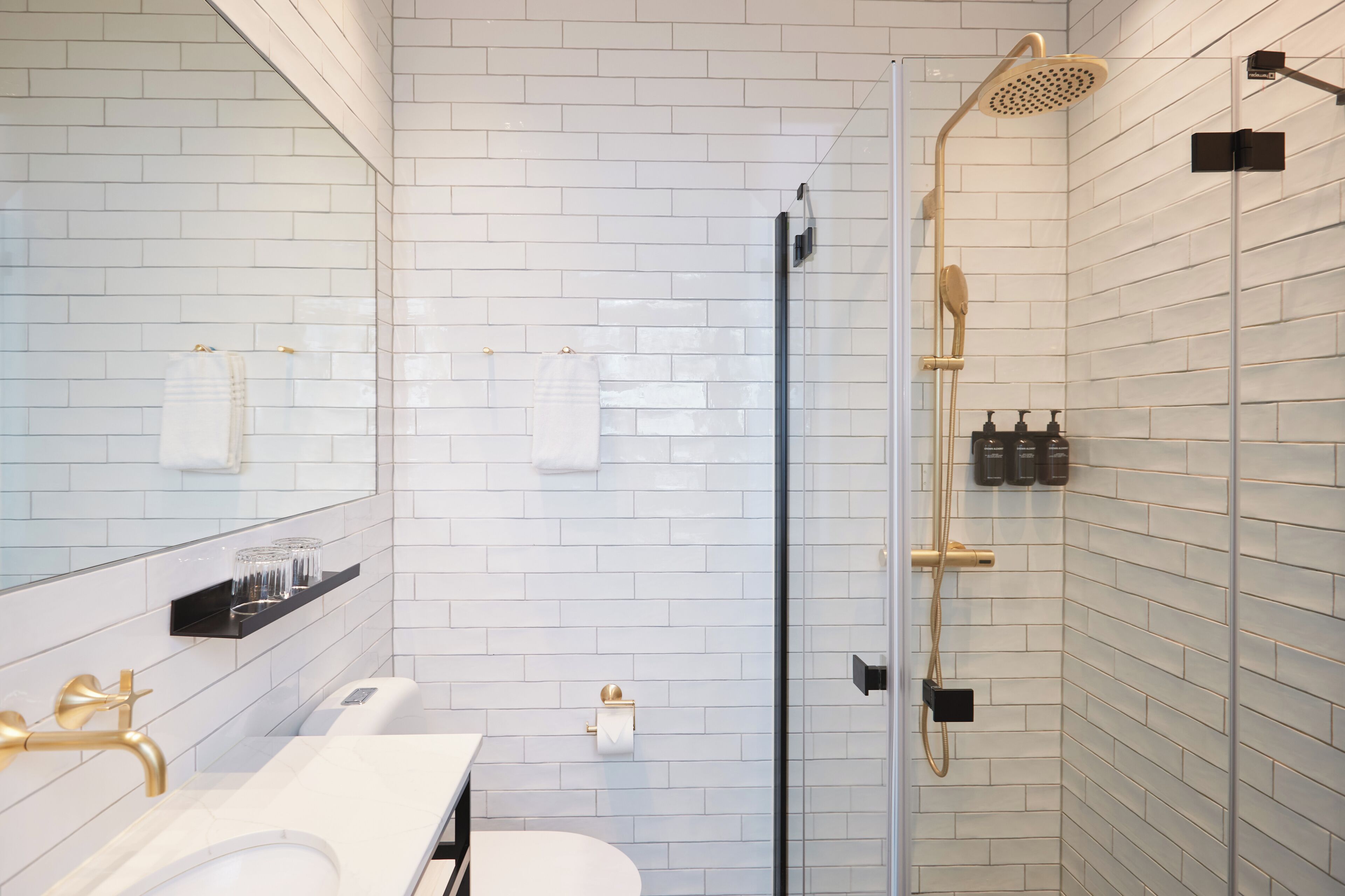 A modern bathroom with white tiled walls, a glass shower enclosure, a white sink, and gold fixtures.