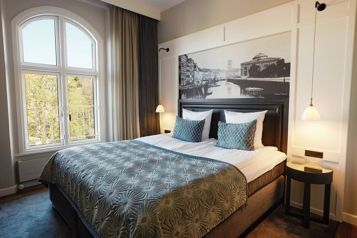 Una habitación de hotel con una cama grande, una ventana con cortinas, un cuadro en la pared y lámparas de noche.