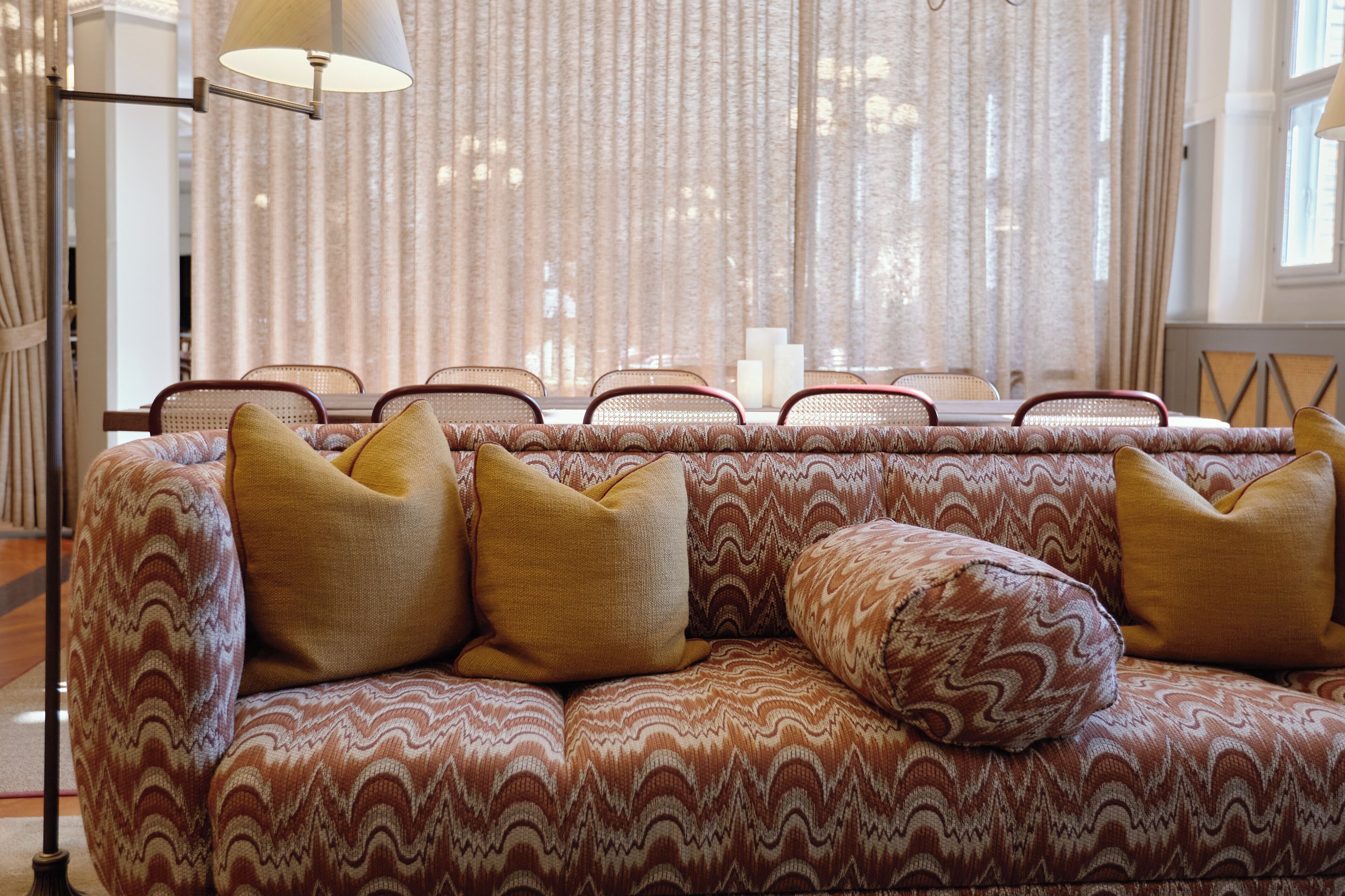 A patterned sofa with cushions, a lamp, and a window with curtains.