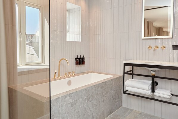 A modern bathroom with a freestanding bathtub, a large mirror, and a built-in shelf with towels. - Ascot Hotel (Copenhagen)