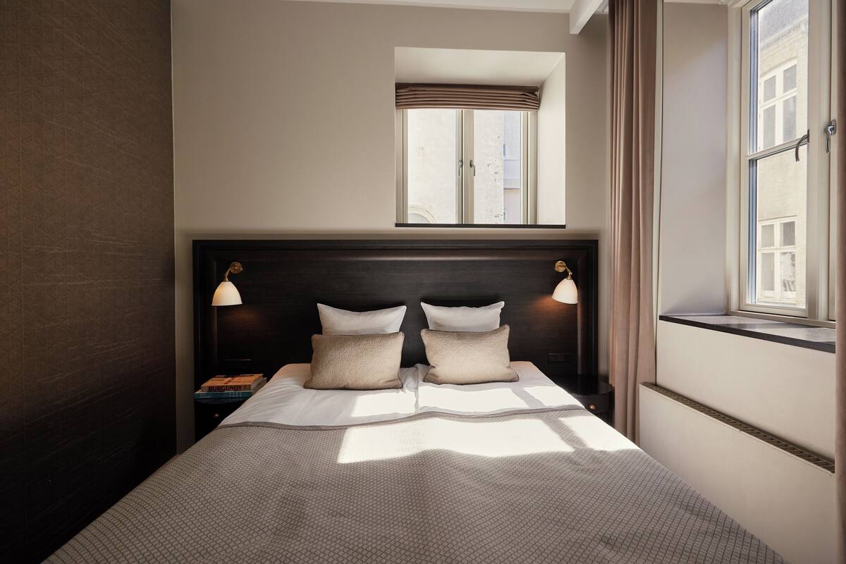 Una cama bien hecha con cabecera, dos lámparas de noche, una mesita de noche y una ventana con cortinas.