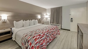 A hotel room with a large bed, two bedside tables with lamps, a nightstand, a desk, and a door. - Red Roof Inn Kingsport (Kingsport)