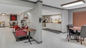 A modern hotel lobby with a Red Roof sign, a seating area with red chairs, a dining area with a wooden table, and a kitchenette in the background. - Red Roof Inn Kingsport (Kingsport)