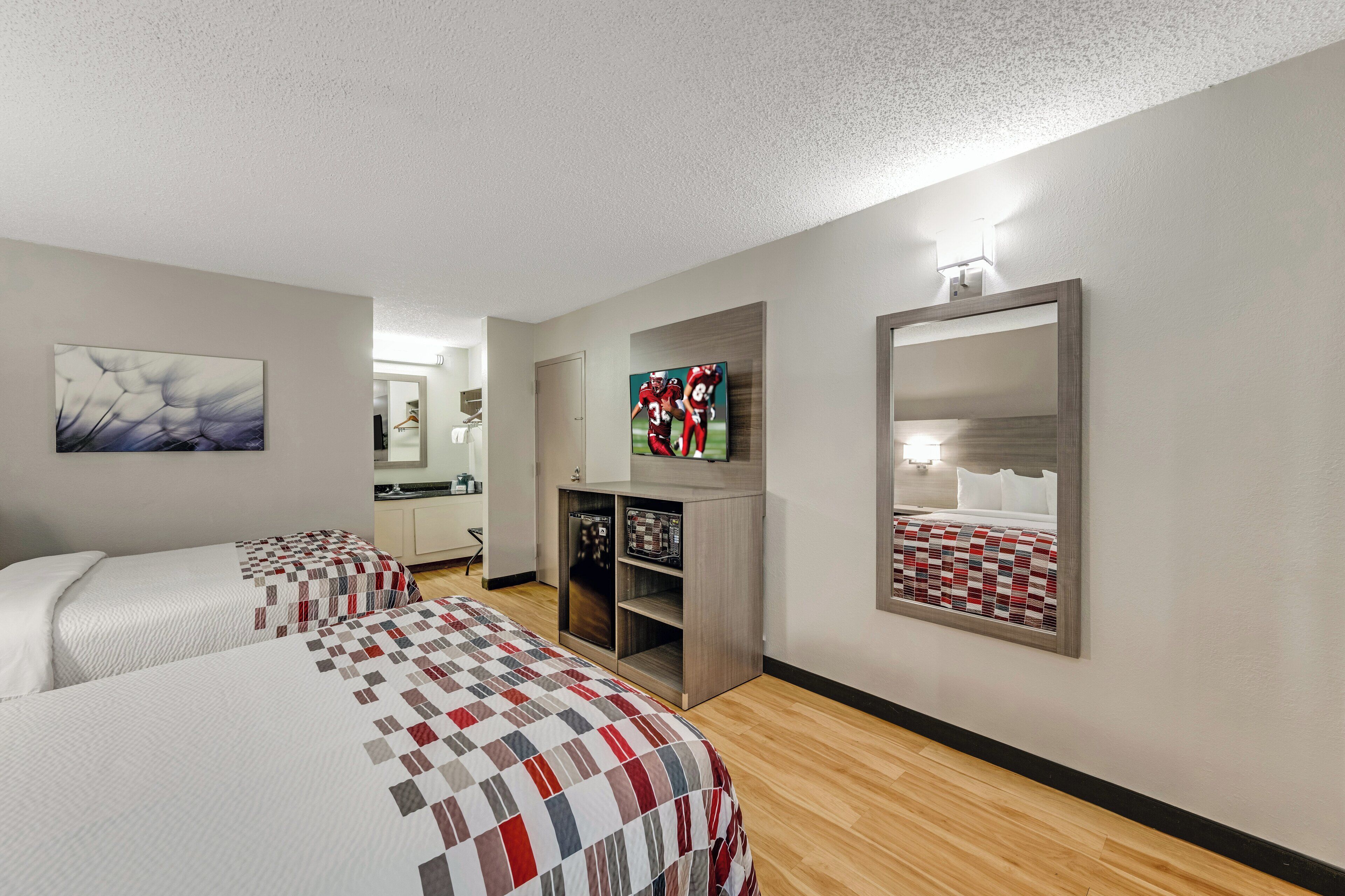 A hotel room with two beds, a TV, a mirror, and a small kitchenette.