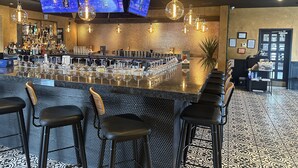 A bar area with a long counter, multiple bar stools, and overhead televisions displaying a cityscape.