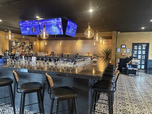 Un espace bar doté d’un long comptoir, de nombreux tabourets et de téléviseurs suspendus au plafond diffusant des images d’un paysage urbain.