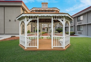 A gazebo with a wooden deck and white railings, surrounded by a well-maintained lawn and a building in the background.