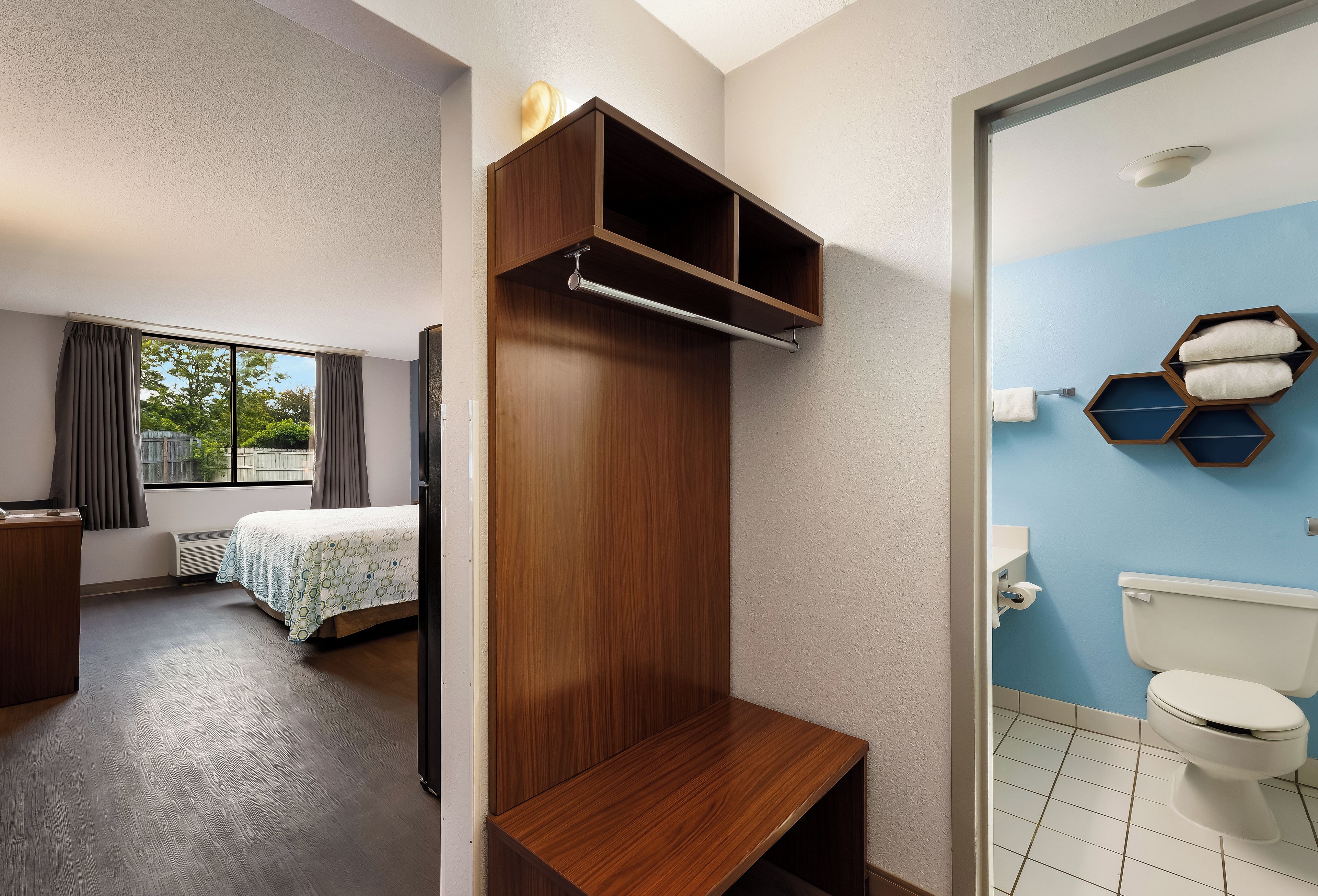 A hotel room with a bed, a wooden wardrobe, a bathroom with a toilet and towel rack, and a window with curtains.