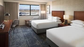 A hotel room with two beds, a desk, a chair, a small table, and a view of an airport.