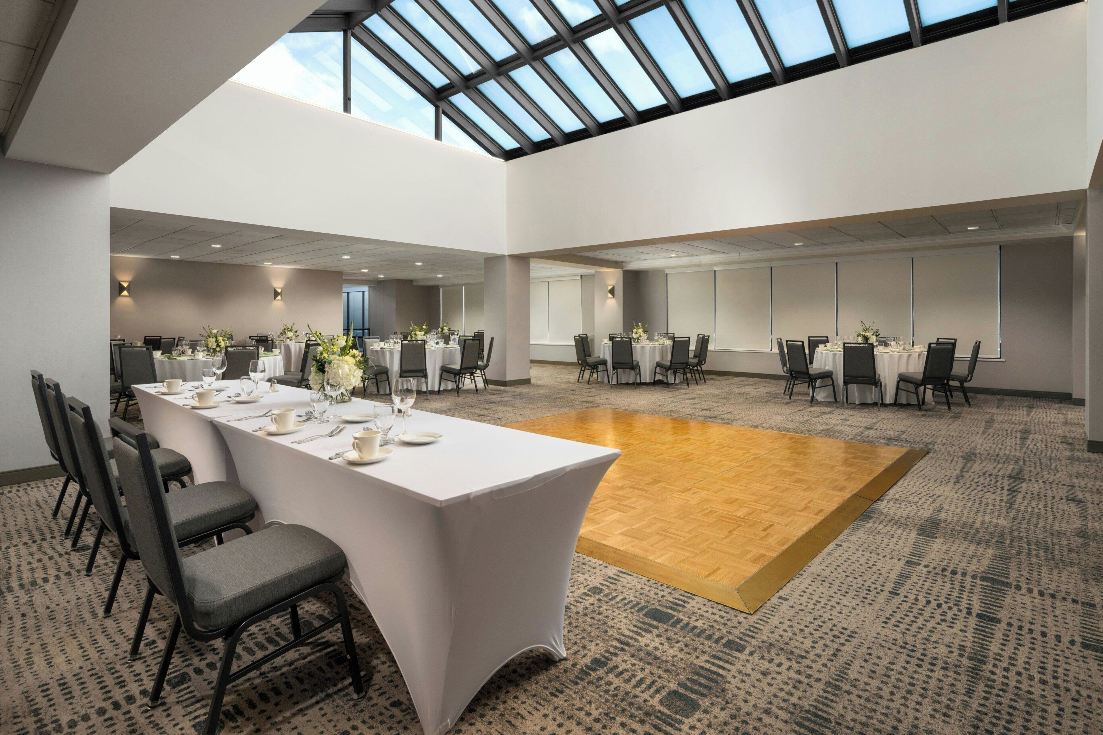Une salle de conférence spacieuse, dotée d’un haut plafond et d’une verrière, comprenant plusieurs tables disposées pour les repas, accompagnées de chaises, une piste de danse centrale et un sol recouvert de moquette.