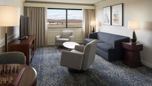Habitación de hotel con un ventanal grande, un sofá, una silla, una mesita, un televisor y un tablero de ajedrez.