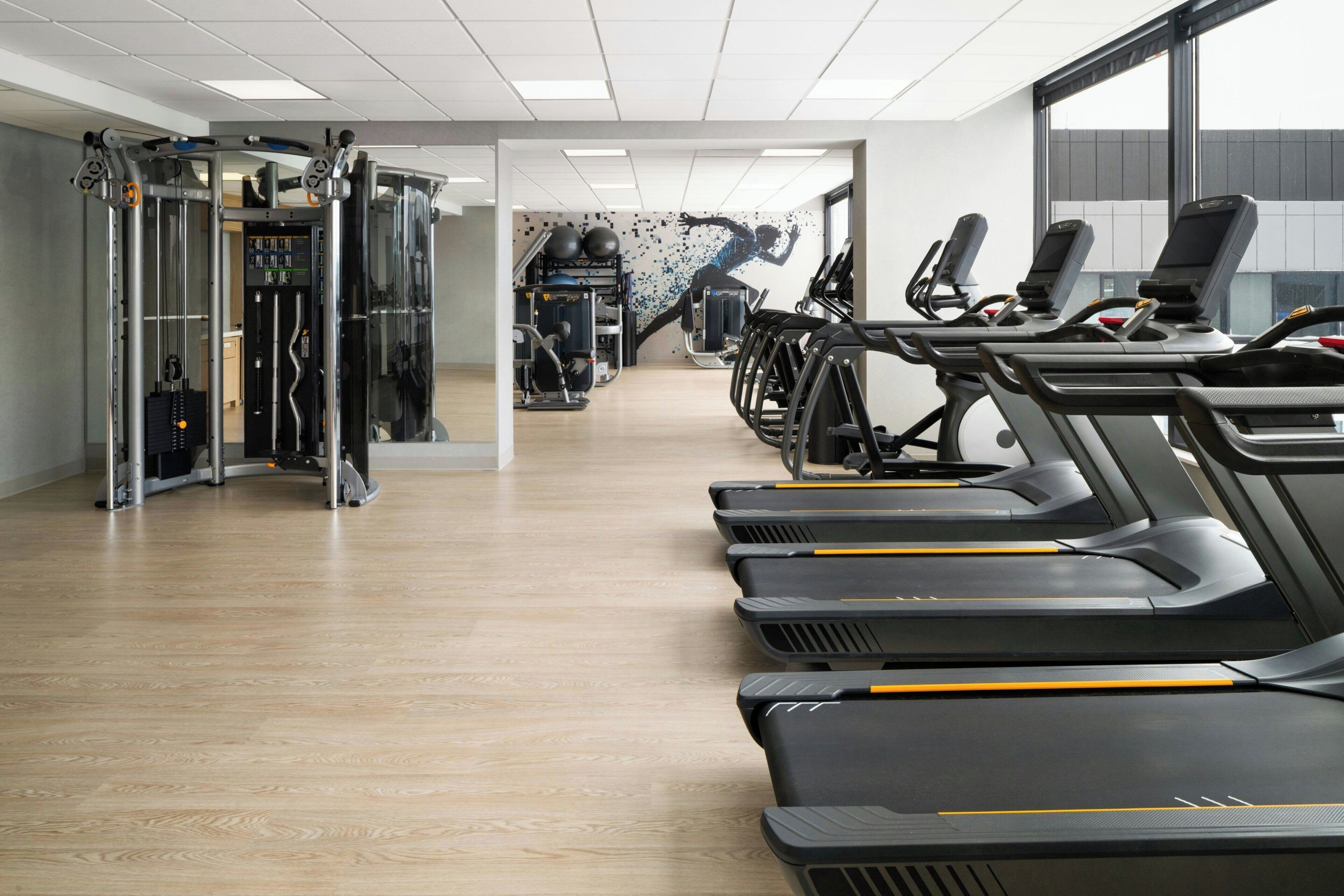 Un gimnasio con cintas de correr, máquinas de pesas y una pared decorada con un mural de temática fitness.
