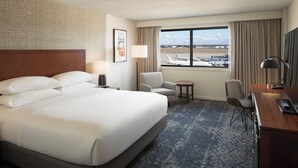Ein Hotelzimmer mit einem großen Bett, einem Schreibtisch, einem Stuhl, einem kleinen Tisch und Blick auf das Flughafengelände.