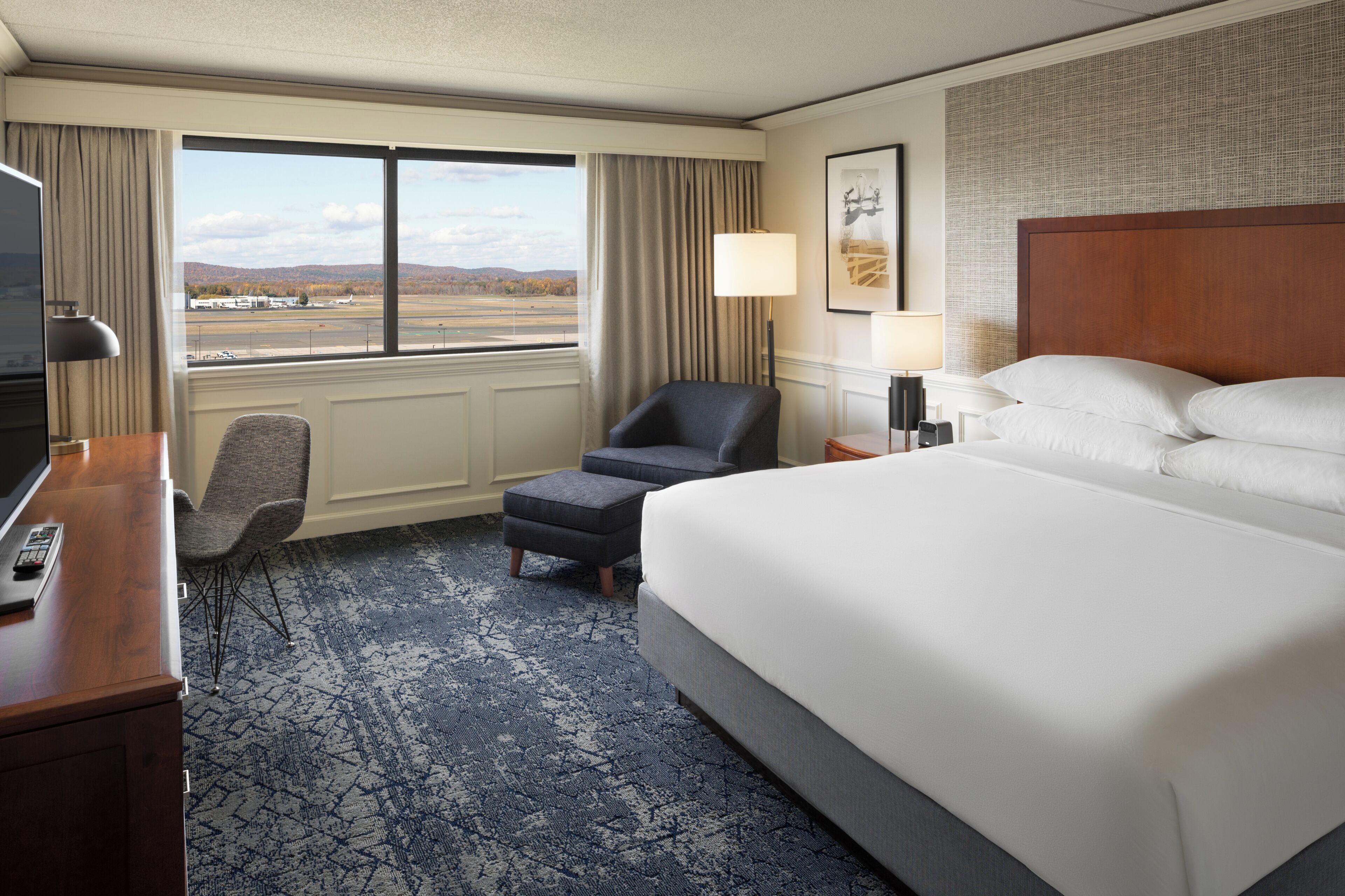 A hotel room with a large bed, a desk with a remote control, a chair, an ottoman, a lamp, and a view of the outdoors.