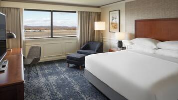 A hotel room with a large bed, a desk with a remote control, a chair, an ottoman, a lamp, and a view of the outdoors.