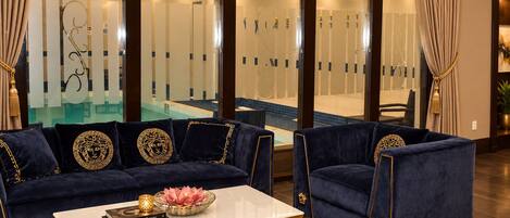 A luxurious living room with a sofa and armchair, a coffee table, and a view of an indoor pool.