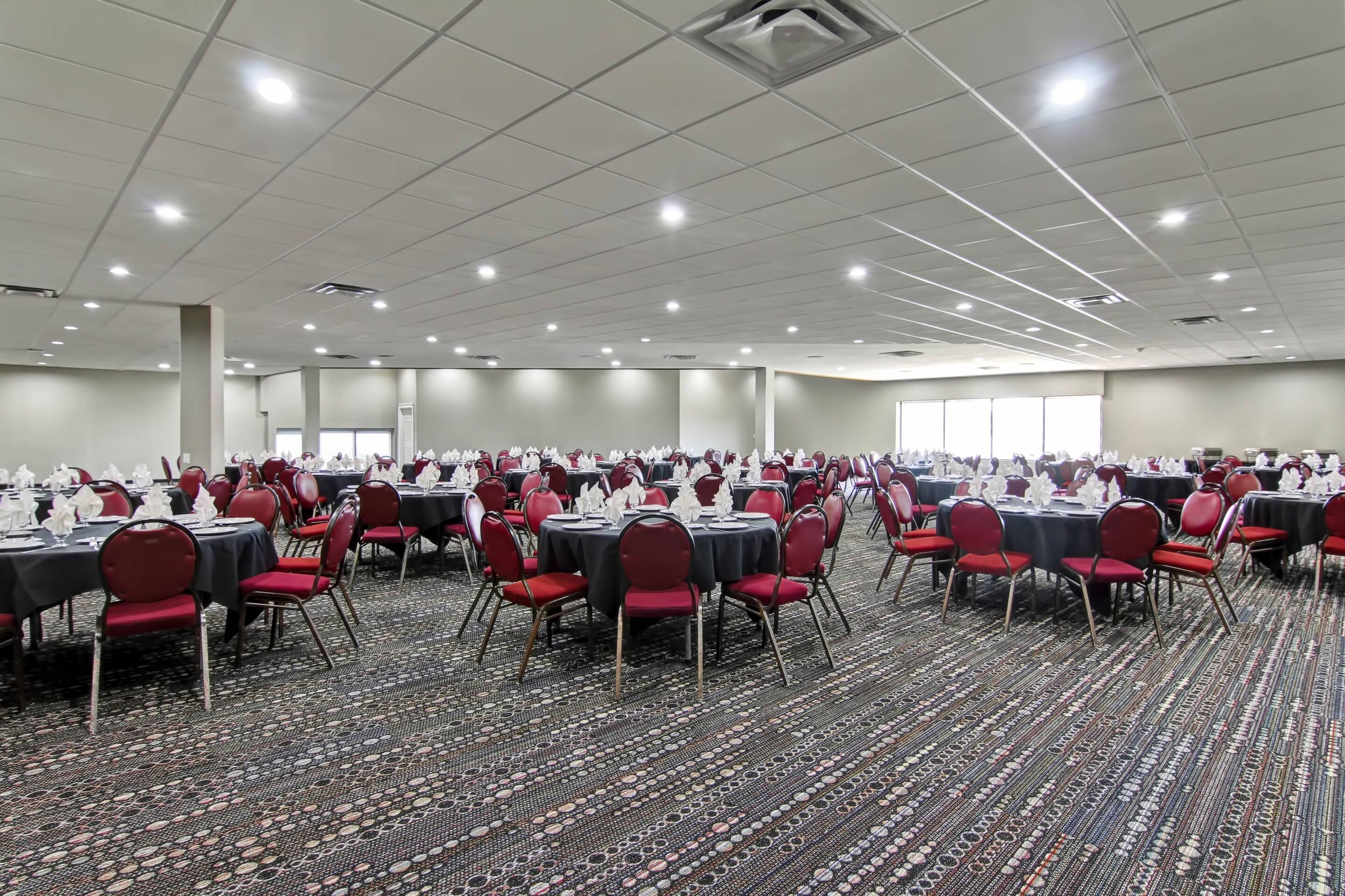 A large banquet hall with rows of round tables, red chairs, and a patterned carpet.