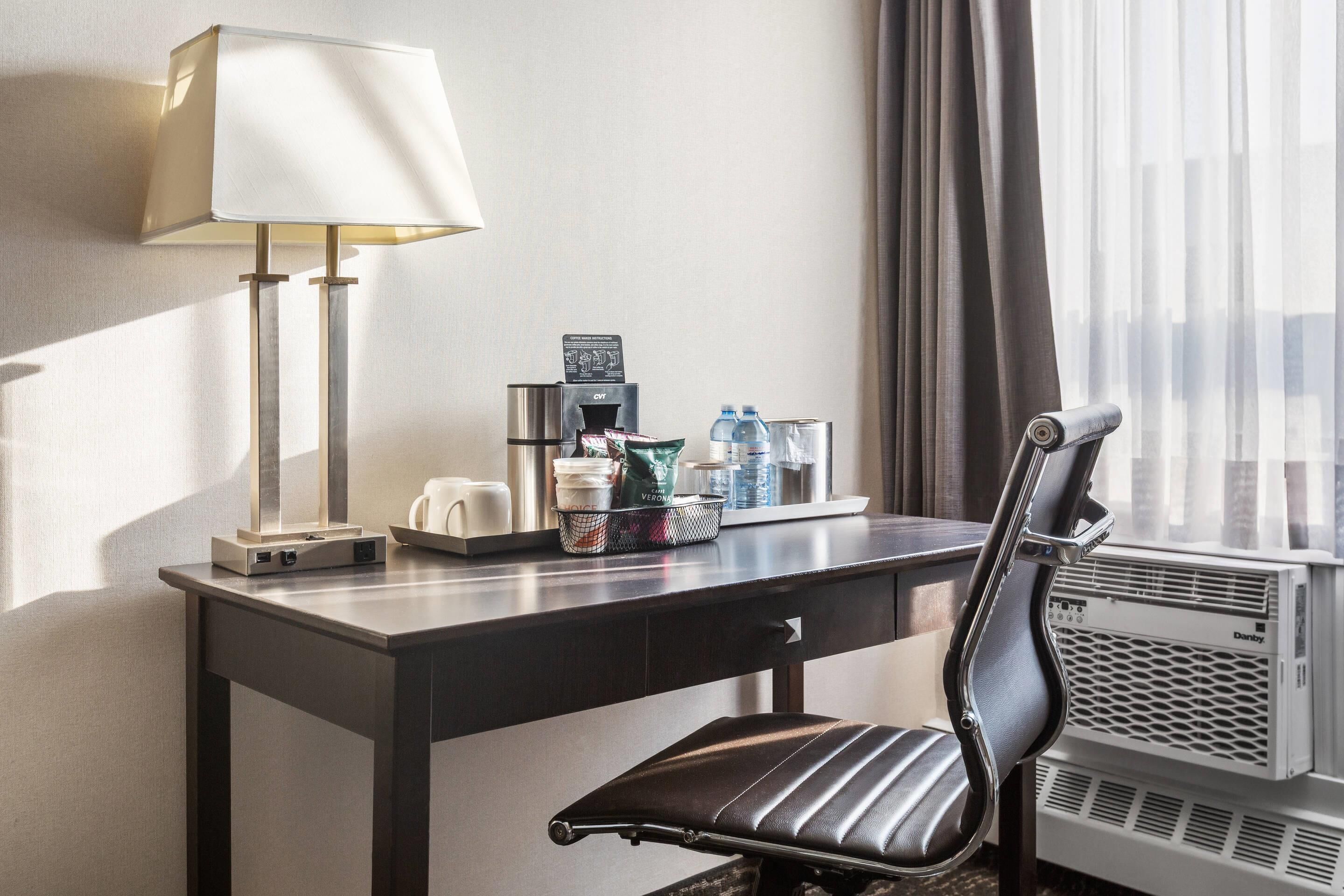 A hotel room with a desk, a chair, a lamp, a coffee maker, a water dispenser, and an air conditioning unit.