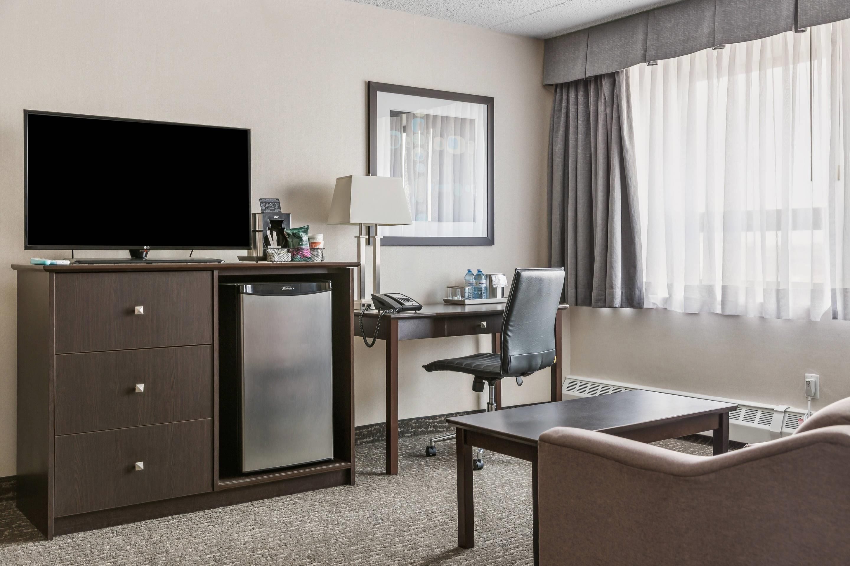 A hotel room with a flat-screen TV, a desk with a chair, a small refrigerator, a bedside table, and a window with curtains.