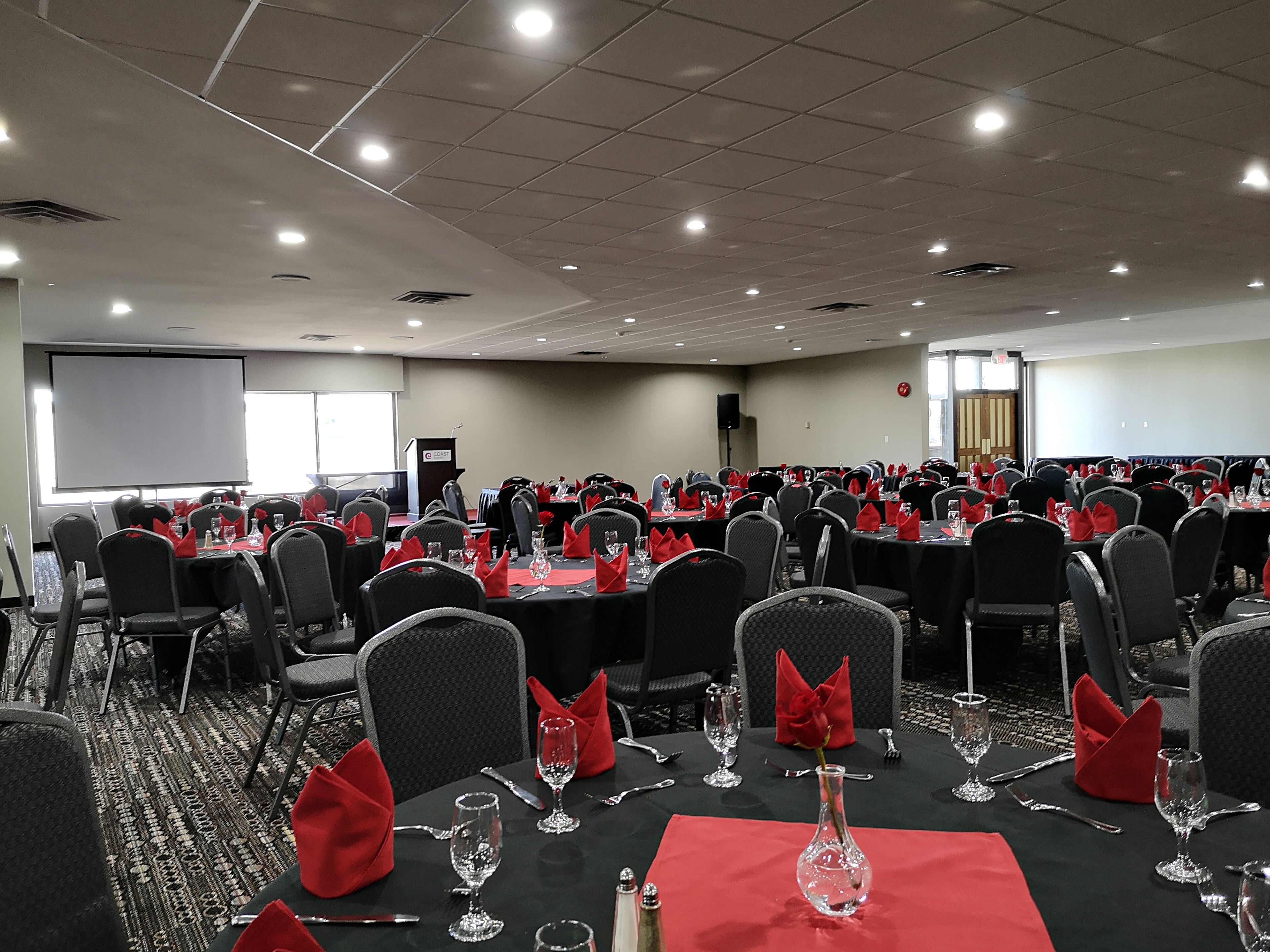 Un salón de conferencias con varias mesas dispuestas para un evento formal, equipado con sillas, mesas y una pantalla de proyector.