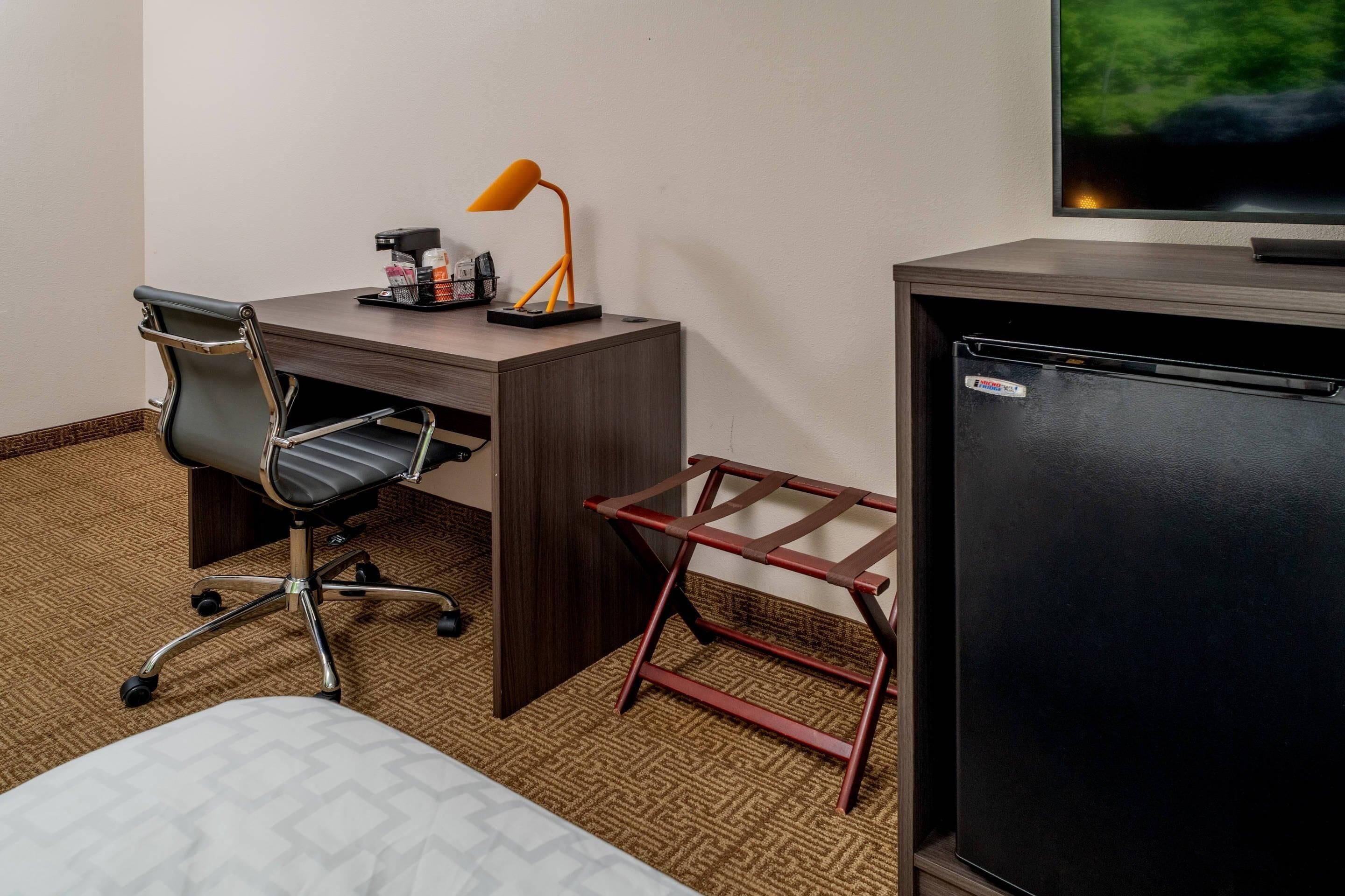 A hotel room with a wooden desk, an office chair, a small table with a lamp, a television, and a refrigerator.