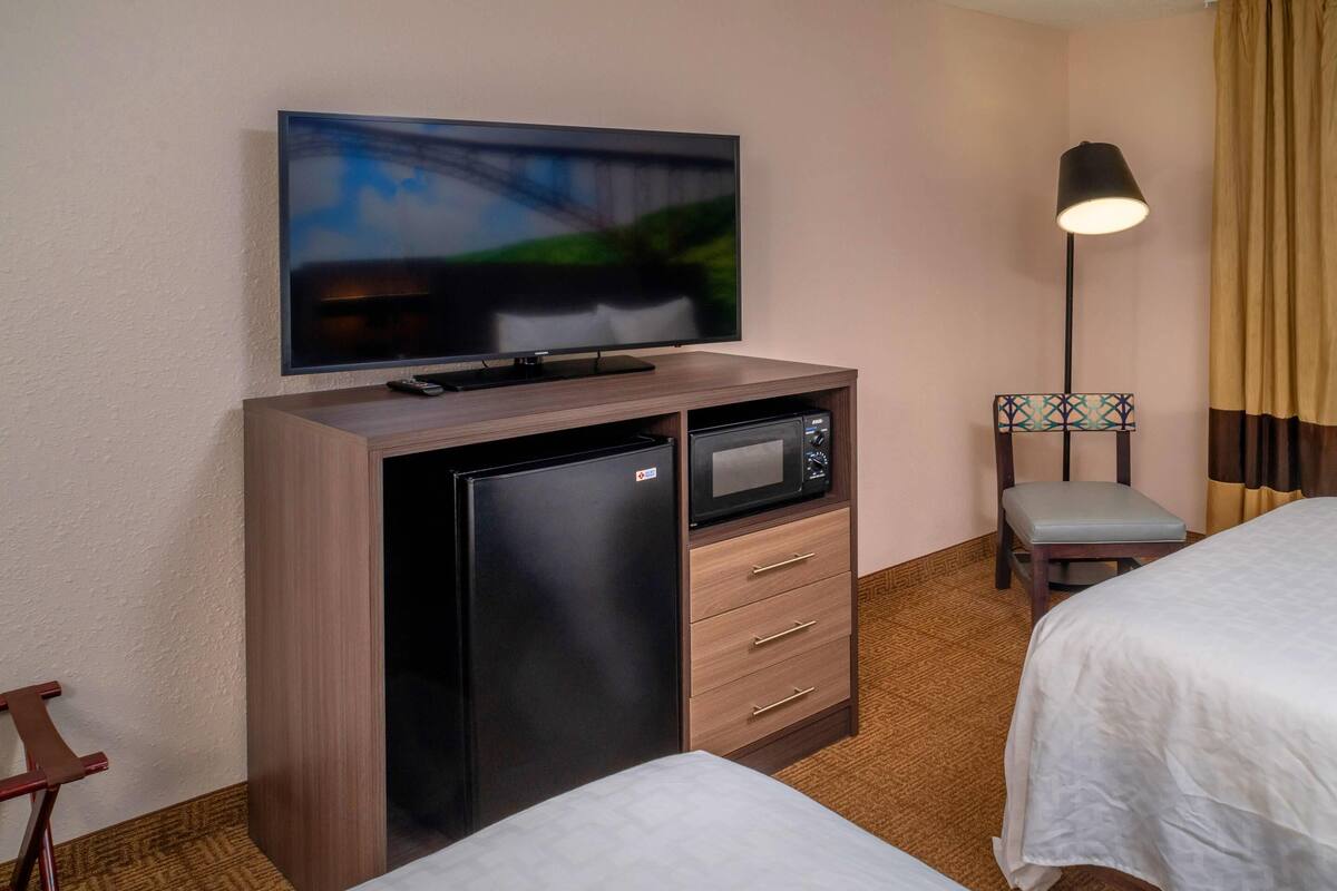 A hotel room with a TV mounted on a wooden cabinet, a microwave, a desk with drawers, a chair, and a bed.